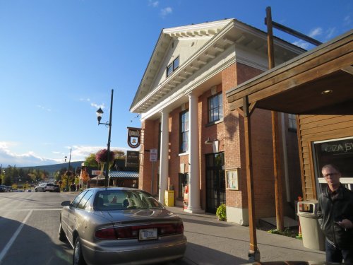 A street view of a brick building with columns, a parked car, and shops under a clear blue sky.