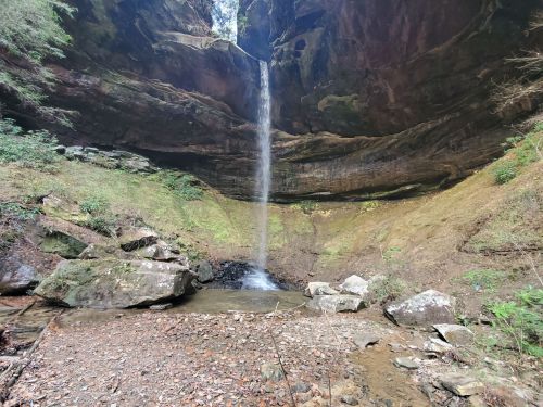 A serene waterfall cascading from a rocky overhang into a small pool, surrounded by lush greenery and rocky terrain.