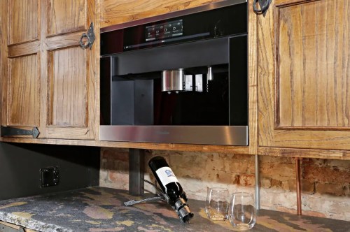 A modern wine dispenser installed in wooden cabinetry, with a bottle of wine and two glasses on the counter below.