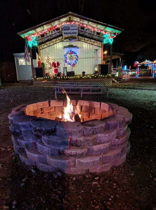 A cozy fire pit in front of a festive stage decorated for the holidays, with Santa and colorful lights in the background.