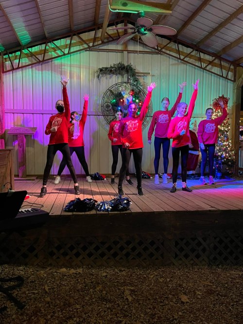 A group of dancers in red shirts performs on stage with colorful lights and holiday decorations in the background.