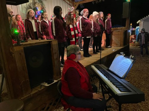 A choir performs on stage while a musician plays piano nearby, all set in a festive outdoor setting.
