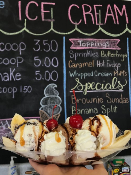A hand holding a banana split topped with cherries, next to a chalkboard menu listing ice cream prices and toppings.