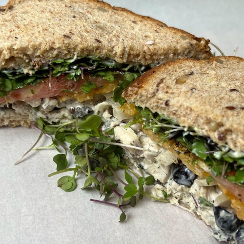 Close-up of a sandwich with layers of vegetables, herbs, and creamy filling, served on whole grain bread.