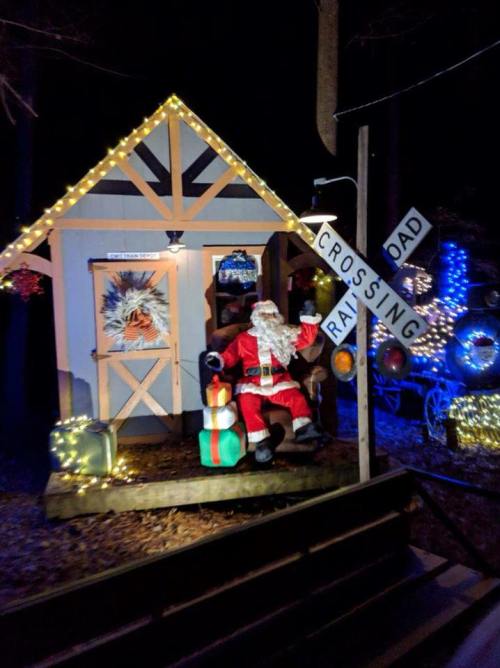 A festive Santa Claus sits on a porch decorated with lights, next to a railroad crossing sign and holiday gifts.
