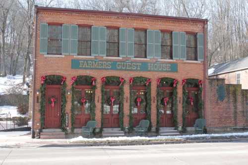 Historic brick building with green shutters, decorated with wreaths, labeled "Farmers' Guest House," in a snowy setting.