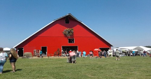 A bright red barn with people gathered outside on a sunny day, surrounded by green grass and tents.