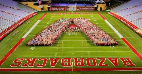 A large group of people forms a giant "A" on a football field, surrounded by stadium seating.