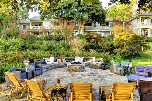A cozy outdoor seating area with a stone fire pit, surrounded by lush greenery and colorful plants.