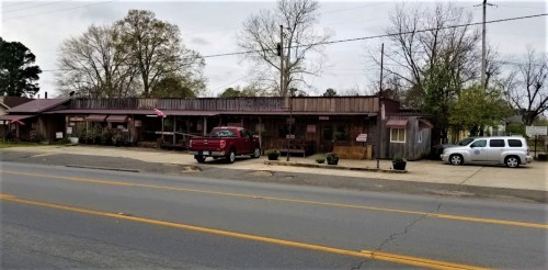 A small town storefront with a red truck parked outside, featuring trees and a cloudy sky in the background.