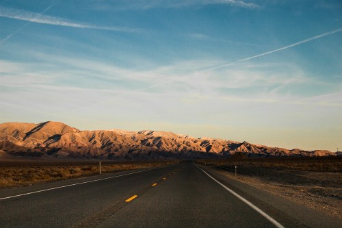 A long, empty road stretches towards mountains under a clear blue sky, with warm sunlight illuminating the landscape.