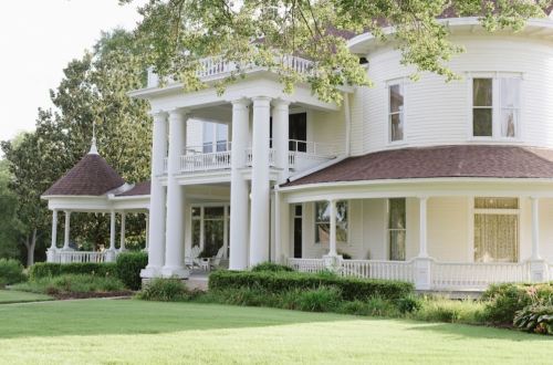 A large, elegant white house with columns, a wraparound porch, and lush green landscaping.