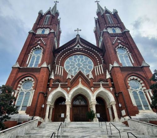 A grand red brick church with two tall spires and a large rose window, set against a cloudy sky.