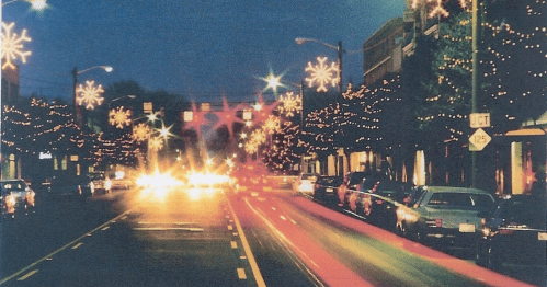A city street at night, decorated with festive lights and snowflake ornaments, with blurred car lights in motion.
