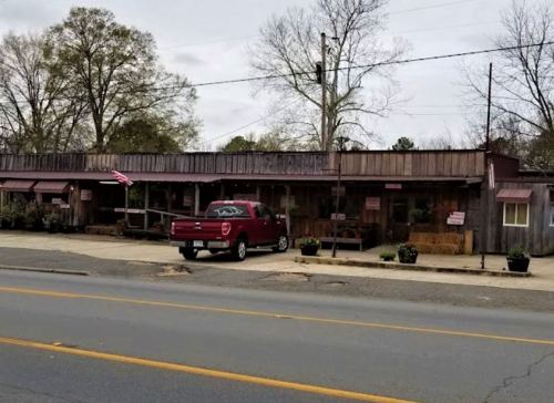 A rustic storefront with a red truck parked outside, featuring potted plants and an American flag.