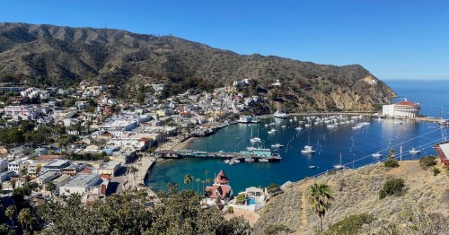 A panoramic view of a coastal town with a marina, hills, and clear blue skies. Boats are docked in the harbor.
