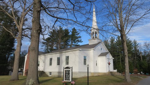 A white church with a tall steeple, surrounded by trees and a clear blue sky, featuring a sign in front.