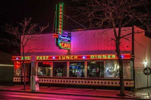 A brightly lit diner at night, featuring neon signs for breakfast, lunch, and dinner. Trees are bare in the foreground.
