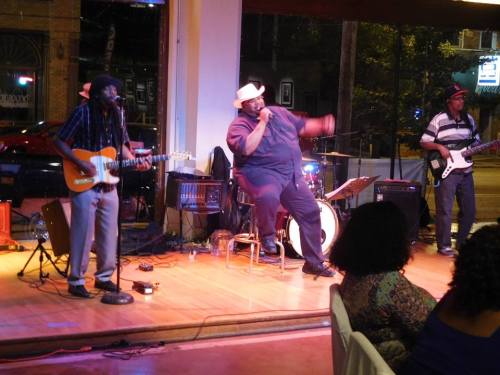 A lively band performs on stage, featuring a guitarist, a seated vocalist, and a bassist, with an audience in view.