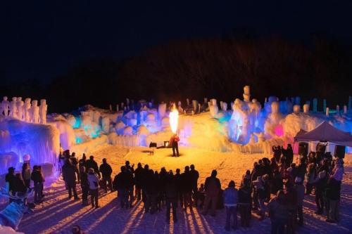 A crowd watches a performer breathing fire in front of colorful ice sculptures at night.