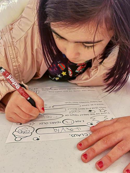 A child with purple hair writes on a worksheet, focused on their task, with colorful designs in the background.