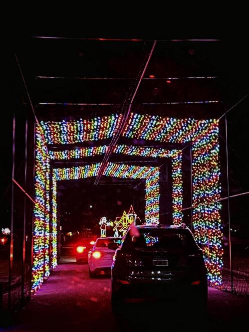 A car drives through a colorful, illuminated tunnel of holiday lights, with festive decorations in the background.