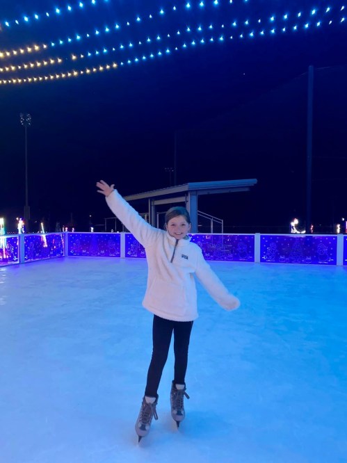 A young girl ice skating on a brightly lit rink, smiling and waving with colorful lights in the background.