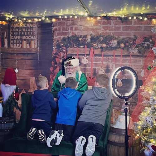 Santa Claus sits on a couch with two children, surrounded by festive decorations and lights.