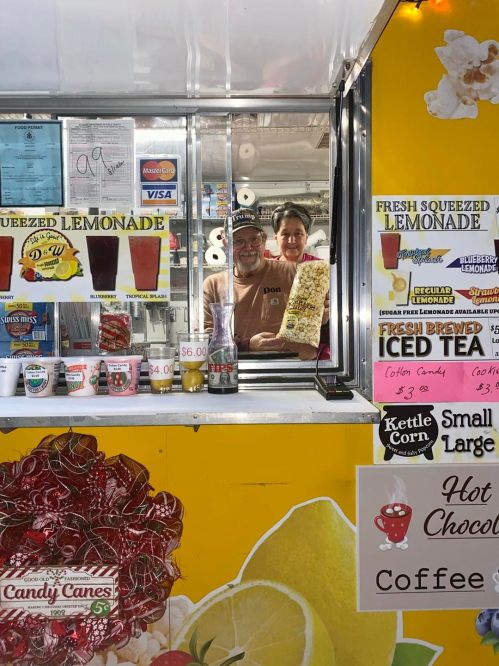 A vendor and a customer smile from a colorful lemonade stand, surrounded by drinks and snacks.