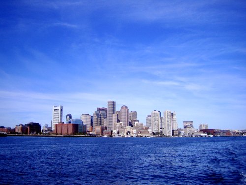 A skyline of tall buildings against a clear blue sky, viewed from across a body of water.
