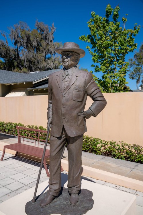 A bronze statue of a man in a suit and hat, standing with a cane against a clear blue sky.