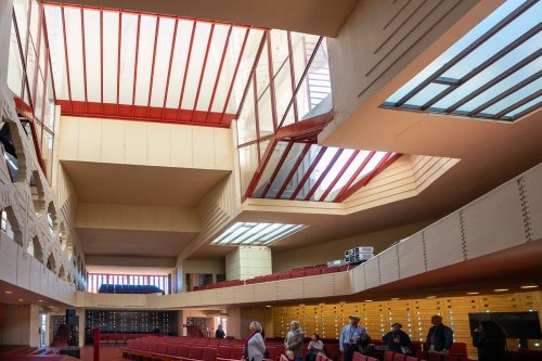 Interior of a modern building with high ceilings, skylights, and red accents, featuring people exploring the space.