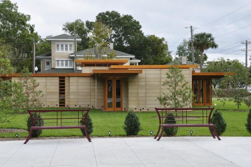 Modern house with wooden accents, surrounded by greenery and benches in a park-like setting.