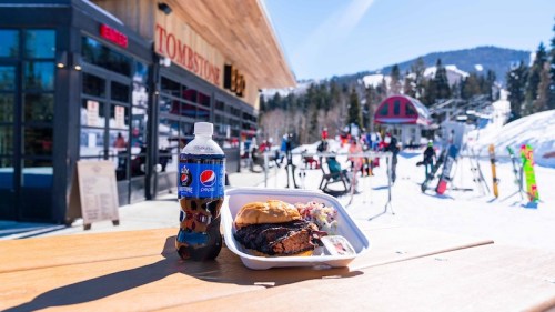 A plate of barbecue chicken and coleslaw with a soda, set on a table outside a ski lodge in a snowy landscape.