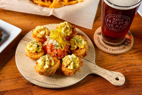 A wooden platter holds six golden, crispy appetizers topped with creamy sauce and garnished, alongside a pint of beer.