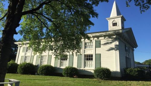 A white building with green shutters and a steeple, surrounded by bushes and trees under a clear blue sky.
