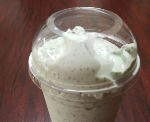 A close-up of a mint-flavored ice cream in a clear cup with a dome lid, sitting on a wooden surface.