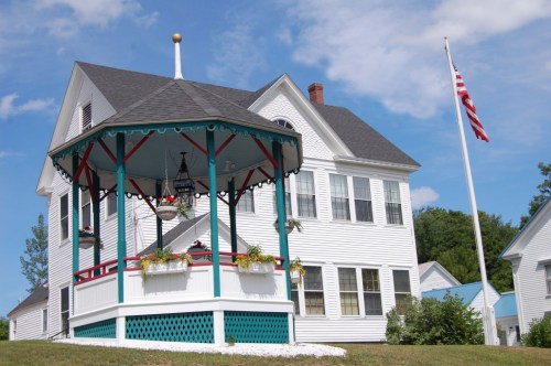 A white house with a gazebo in front, surrounded by greenery and a flagpole displaying the American flag.