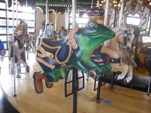 A colorful frog carousel figure in motion, surrounded by traditional horse figures on a carousel.