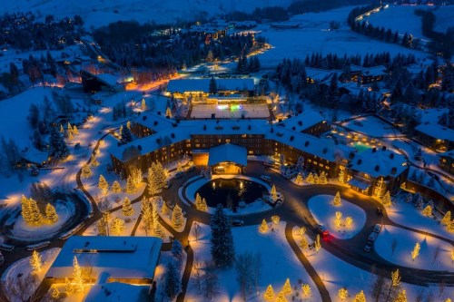 Aerial view of a snow-covered resort illuminated with warm lights, surrounded by trees and mountains at night.