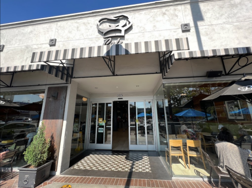 Exterior of a café with a striped awning, glass doors, and outdoor seating under umbrellas on a sunny day.