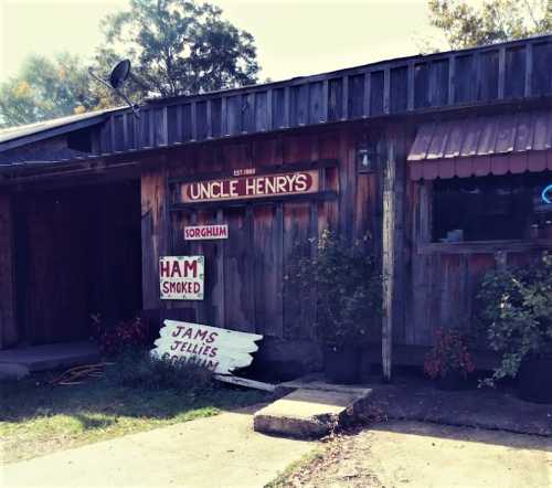 A rustic wooden building with signs for "Uncle Henry's," "Sorghum," "Ham Smoked," and "Jams Jellies."