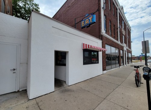 A small white building with a red-striped awning and a sign that says "EAT," next to a brick building and a bicycle.