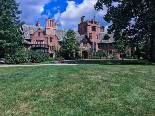 A large, historic brick mansion surrounded by lush greenery and a clear blue sky.