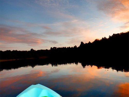 A serene sunset over a calm lake, with colorful reflections and silhouettes of trees in the background.