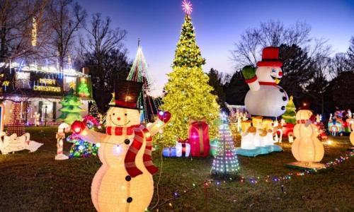 Colorful Christmas decorations with inflatable snowmen, a lit tree, and festive lights in a yard at dusk.