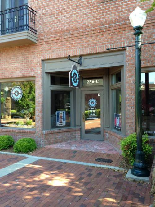 A brick storefront with large windows and a sign reading "236-C," surrounded by greenery and a lamp post.