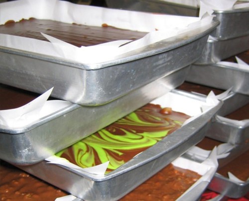 Three stacked metal trays filled with different types of chocolate and swirled green treats, lined with parchment paper.