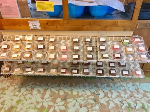 A display of assorted fudge samples in clear containers on a wire rack, with labels for each flavor.
