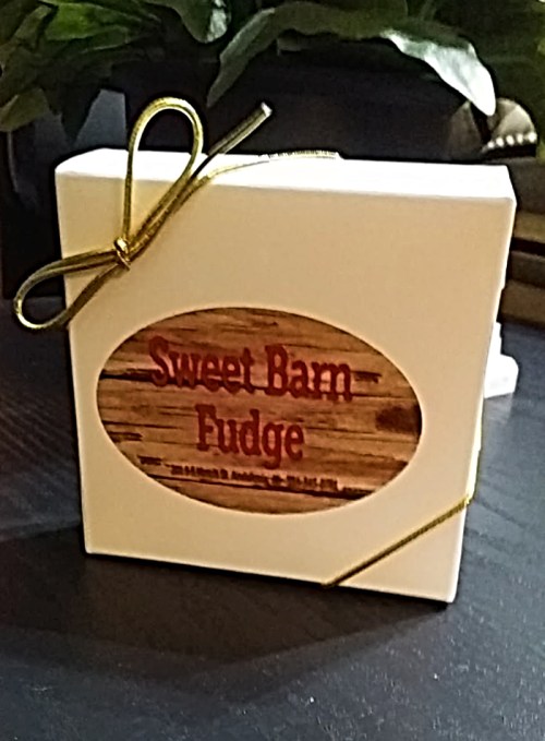 A white gift box tied with a gold ribbon, featuring a label that reads "Sweet Barn Fudge" on a wooden background.